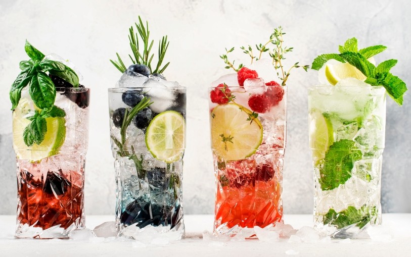 Cocktails or mocktails cold drinks. Classic summer refreshing long drink in highballs with berries, lime, herbs and ice on gray bar counter background